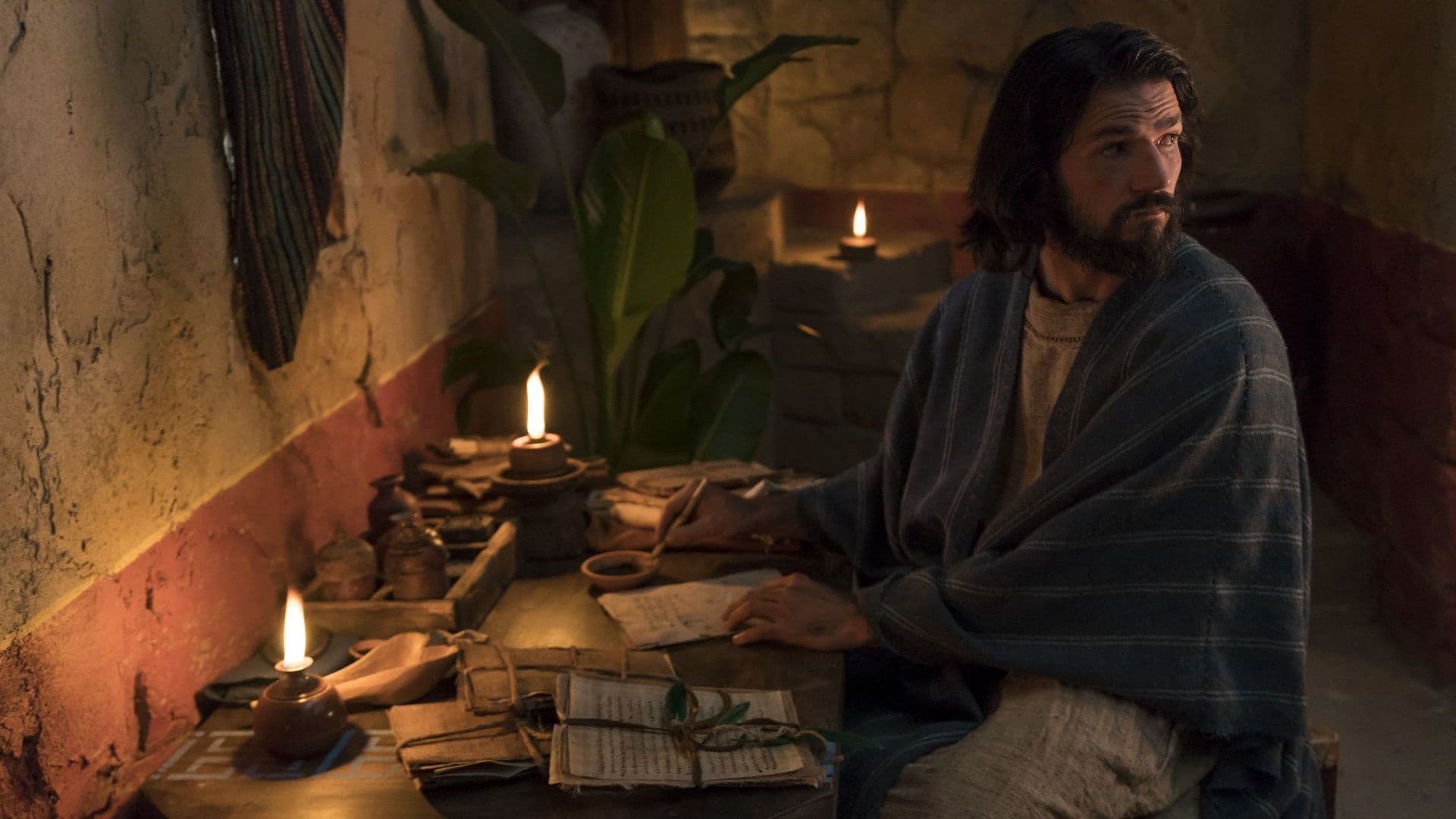 Alma the Younger writes on parchment as he sits at a table lit with candles.